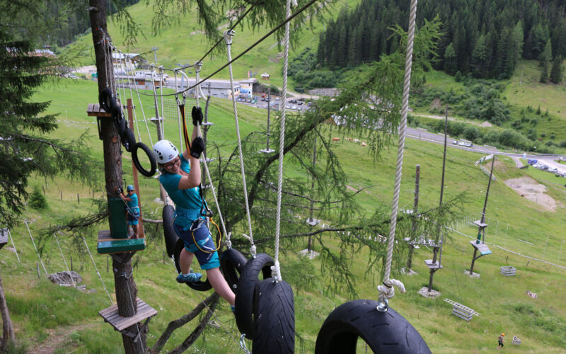 Eine Person mit Sicherheitsausrüstung navigiert durch einen Hochseilgarten, hält sich an Seilen fest und tritt auf aufgehängte Reifen, während im Hintergrund eine Graslandschaft und Bäume zu sehen sind.