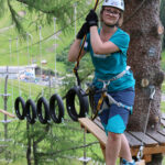 Eine Person mit Sicherheitsausrüstung steht auf einer Holzplattform in einem Hochseilgarten und hält sich an einem Seil fest, das an einem Gurt befestigt ist.