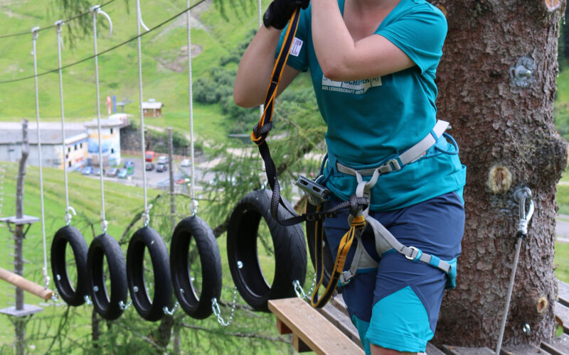Eine Person mit Sicherheitsausrüstung steht auf einer Holzplattform in einem Hochseilgarten und hält sich an einem Seil fest, das an einem Gurt befestigt ist.