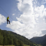 Eine Person, die einen Helm und ein Gurtzeug trägt, fährt auf einer Seilrutsche hoch über einem bewaldeten Tal mit Bergen und einem teilweise bewölkten Himmel im Hintergrund.