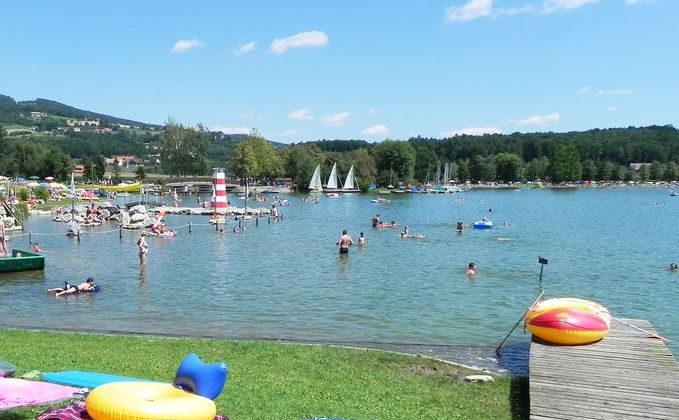 Menschen schwimmen und entspannen sich an einem sonnigen Tag an einem Strand am See, mit Segelbooten im Hintergrund und Schlauchbooten auf dem Rasen in der Nähe des Wassers.