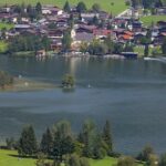 Eine kleine Stadt mit roten Dächern und einer Kirche liegt am Rande eines Sees, umgeben von grünen Feldern und Bäumen.