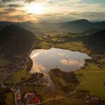 Luftaufnahme eines von Dörfern, grünen Feldern und Hügeln umgebenen Sees bei Sonnenuntergang, mit Wolken und Bergen im Hintergrund.