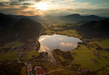 Luftaufnahme eines von Dörfern, grünen Feldern und Hügeln umgebenen Sees bei Sonnenuntergang, mit Wolken und Bergen im Hintergrund.
