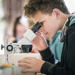 Ein Junge untersucht an einem Tisch eine Probe unter dem Mikroskop. Im Hintergrund sind unscharfe Menschen und eine Landschaft im Freien zu sehen.