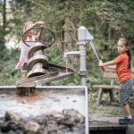 Zwei Kinder interagieren mit einer Wasserspielanlage im Freien. Das eine dreht eine Handpumpe, das andere bedient eine Archimedische Schraube. Im Hintergrund sind eine Holzbank und Bäume zu sehen.