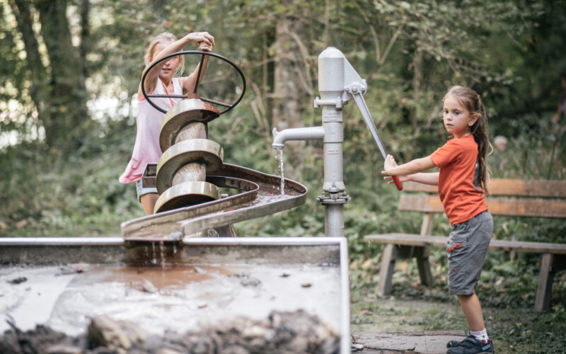 Zwei Kinder interagieren mit einer Wasserspielanlage im Freien. Das eine dreht eine Handpumpe, das andere bedient eine Archimedische Schraube. Im Hintergrund sind eine Holzbank und Bäume zu sehen.