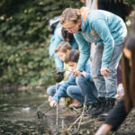 Mehrere Kinder hocken an einem Teich und verwenden Netze, um Gegenstände im Wasser zu sammeln oder zu beobachten, während sie von Grün umgeben sind.