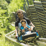 Ein Erwachsener und ein Kind fahren an einem sonnigen Tag gemeinsam auf einer von Grün umgebenen Alpenachterbahn.