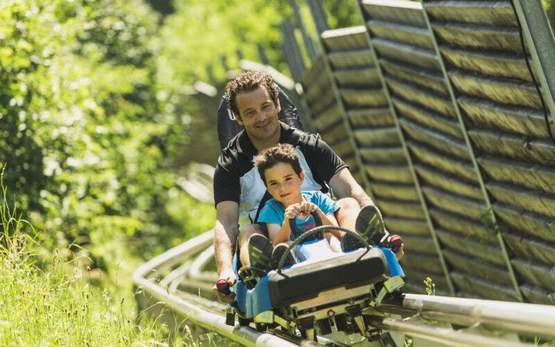 Ein Erwachsener und ein Kind fahren an einem sonnigen Tag gemeinsam auf einer von Grün umgebenen Alpenachterbahn.