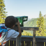 Ein Kind mit einem blauen Rucksack blickt durch einen grünen Landschaftsbetrachter auf eine von Bäumen umgebene Berglandschaft.