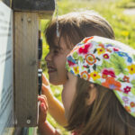 Zwei Kinder schauen sich an einem sonnigen Tag eine Informationstafel im Freien genau an; das eine trägt einen bunten Blumenhut, das andere blinzelt zum Lesen.
