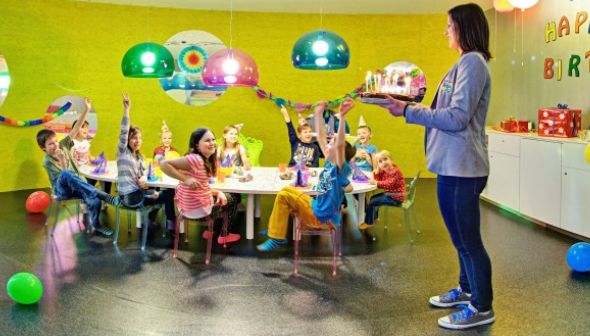 Eine Frau hält eine Geburtstagstorte in der Hand, während Kinder in einem bunt geschmückten Raum um einen Tisch mit Partyhüten und Dekorationen sitzen und ihre Hände heben.