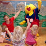 Eine Gruppe kleiner Kinder und ein Erwachsener sitzen auf dem Boden und heben die Arme, während eine Person in einem gelben Zeichentrickfilmkostüm in der Nähe in einem dekorierten Raum steht.