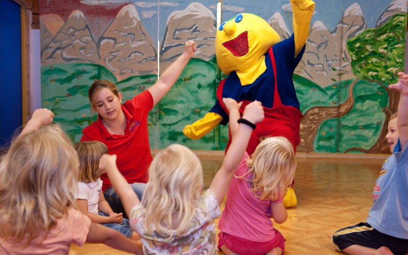 Eine Gruppe kleiner Kinder und ein Erwachsener sitzen auf dem Boden und heben die Arme, während eine Person in einem gelben Zeichentrickfilmkostüm in der Nähe in einem dekorierten Raum steht.