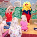 Eine Gruppe kleiner Kinder sitzt auf dem Boden und sieht sich einem Erwachsenen und einer Person in einem gelben Kostüm gegenüber, die in einem farbenfrohen Raum an einer interaktiven Aktivität teilnehmen.