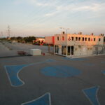 Leerer Parkplatz mit blauen Markierungen, ein kleines zweistöckiges Modulgebäude mit Beschilderung und ein paar Busse und Container im Hintergrund, unter Stromleitungen bei Sonnenuntergang.