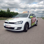 Ein weißer Volkswagen mit dem Schriftzug "sauer" und einem Schild für Fahrschüler parkt auf einer asphaltierten Straße unter einem bewölkten Himmel.