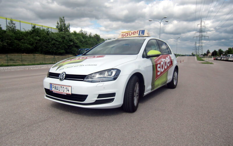 Ein weißer Volkswagen mit dem Schriftzug "sauer" und einem Schild für Fahrschüler parkt auf einer asphaltierten Straße unter einem bewölkten Himmel.