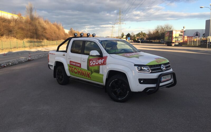 Ein weißer Volkswagen Amarok Pickup mit dem Schriftzug Sauer Technik" ist auf einer leeren asphaltierten Straße in der Nähe eines Industriegebiets geparkt.