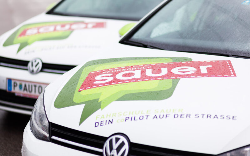 Zwei weiße Volkswagen mit den Logos "Fahrschule Sauer" und "Dein CoPilot auf der Straße" auf den Motorhauben, nebeneinander auf einer nassen Fläche geparkt.