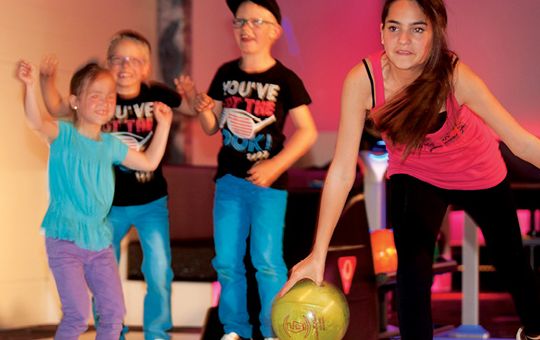 Ein Mädchen kegelt mit einer grünen Kugel, während drei Kinder hinter ihr in einer hell erleuchteten Kegelbahn jubeln und zusehen.