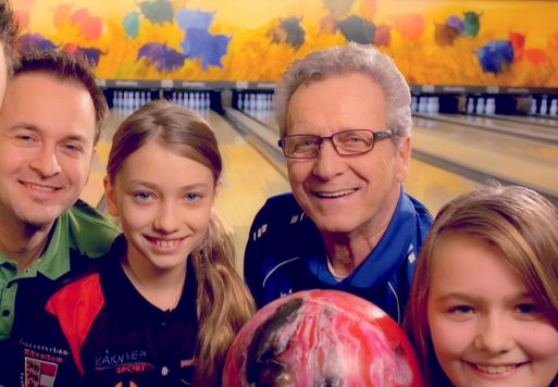Vier Personen, darunter zwei Mädchen und zwei Männer, lächeln gemeinsam auf einer Bowlingbahn und halten eine Bowlingkugel mit bunten Pins und einem gemalten Wandbild im Hintergrund.