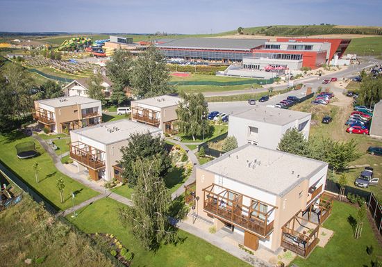 Luftaufnahme von mehreren modernen zweistöckigen Häusern mit Balkonen, grünen Rasenflächen und einem großen Gebäudekomplex mit Parkplätzen im Hintergrund.