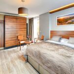 Modernes Hotelzimmer mit großem Bett, Holzmöbeln, Schreibtisch, an der Wand montiertem Fernseher, kleinem Tisch mit zwei Stühlen und einem Landschaftsgemälde über dem Bett.