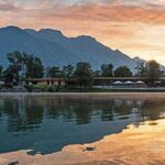 In einem ruhigen See spiegeln sich Bäume, ein Gebäude und Berge bei Sonnenuntergang, mit orangefarbenen Wolken am Himmel darüber.