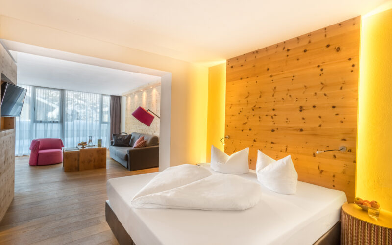 Modernes Hotelzimmer mit einem Doppelbett, einer Akzentwand aus Holz und einer separaten Sitzecke mit einem Sofa, einem rosa Sessel und einem großen Fenster mit Tageslicht.