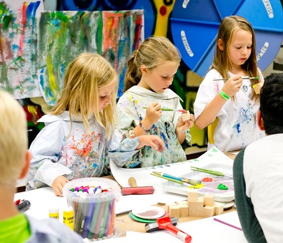 Drei kleine Kinder in mit Farbe bespritzten Kitteln konzentrieren sich auf das Malen und Zeichnen an einem Tisch in einem Kunstunterrichtsraum, umgeben von Kunstzubehör.