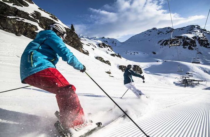 Zwei Personen fahren eine frisch präparierte Piste in einer verschneiten Berglandschaft mit Skiliften und felsigen Gipfeln im Hintergrund hinunter.