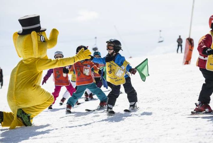 Eine Person in einem gelben Elefantenkostüm begrüßt eine Gruppe von Kindern, die auf einer verschneiten Piste das Skifahren lernen.