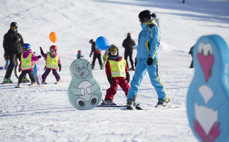 Kinder und ein Skilehrer fahren auf einer verschneiten Piste Ski. Sie tragen Helme und bunte Westen, im Hintergrund sind Cartoon-Tierausschnitte und Luftballons zu sehen.