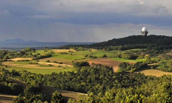 Wogende grüne und braune Felder mit Baumgruppen, ein bewaldeter Hügel und eine große weiße Radarkuppel, die auf dem Hügel unter einem bewölkten Himmel zu sehen ist.
