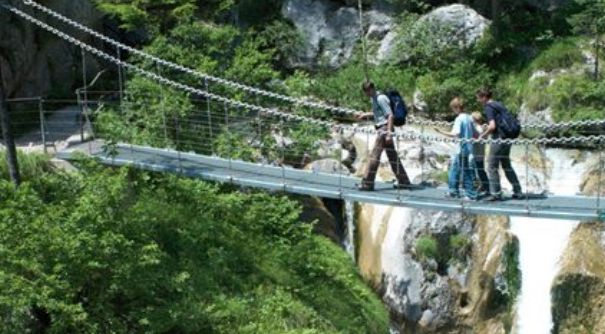Drei Personen überqueren eine schmale Hängebrücke über eine felsige Schlucht mit Grünpflanzen und einem Wasserfall im Hintergrund.