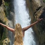 Eine Person mit langem blondem Haar steht mit ausgestreckten Armen vor einem Wasserfall, der über felsiges Gelände stürzt.