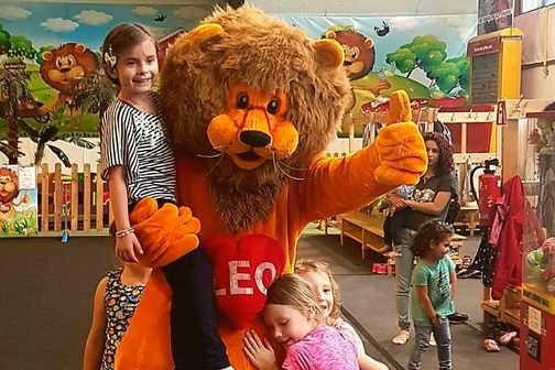 Eine Person in einem Löwenmaskottchenkostüm mit der Aufschrift "LEO" posiert mit lächelnden Kindern in einem mit Cartoon-Löwen dekorierten Indoor-Spielbereich.