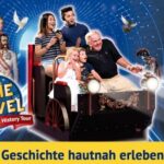Eine Gruppe von Personen auf einem Fahrgeschäft schaut aufgeregt auf historische Figuren, die sie umgeben; der Text lautet: "TIME TRAVEL Magic Vienna History Tour" und "Geschichte hautnah erleben!.