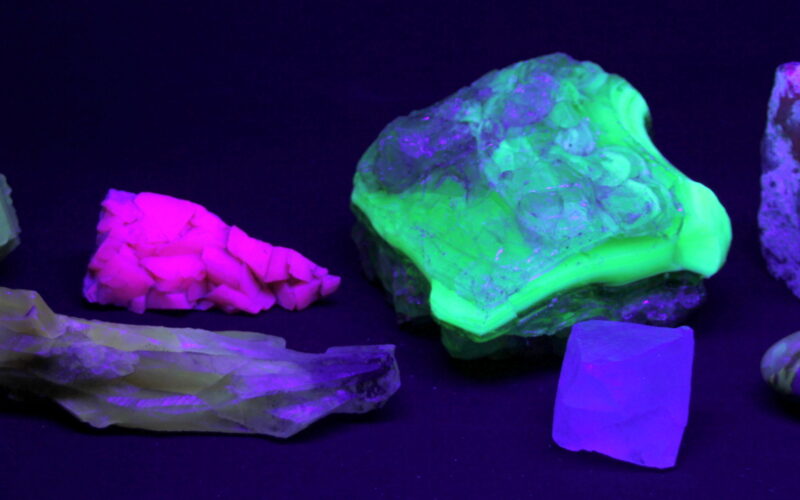 Eine Sammlung von Mineralien, die unter ultraviolettem Licht in verschiedenen Neonfarben leuchten, darunter leuchtend grüne, rosa und violette Farbtöne.