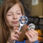 Ein Mädchen untersucht eine Münze mit einem Vergrößerungsglas, wobei es die Münze in der einen und die Lupe in der anderen Hand hält, vor einem unscharfen Hintergrund.