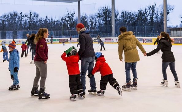 Menschen verschiedenen Alters beim Schlittschuhlaufen in einer Eishalle, einige halten sich an den Händen, andere laufen unabhängig voneinander. Draußen sind Bäume und ein bewölkter Himmel durch Glasscheiben zu sehen.