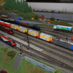 Modelleisenbahnanlage mit mehreren Gleisen, verschiedenen Modelleisenbahnen, Güterwagen und einer kleinen Grünanlage mit Bäumen und Miniaturgebäuden im Hintergrund.