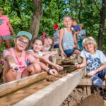 Eine Gruppe Kinder durchsucht im Freien lächelnd eine Holzschleuse und ist unter den Bäumen mit einer praktischen Aktivität beschäftigt.