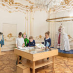 Drei Kinder sitzen an einem Zeichentisch in einem Museumsraum mit verschnörkelten Wänden, ausgestellten historischen Kostümen und einem Kronleuchter über dem Kopf.