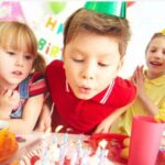 Drei Kinder mit Partyhüten sitzen an einem Tisch, während der Junge in der Mitte die Kerzen auf einer Geburtstagstorte auspustet.