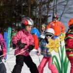 Kinder in bunten Skiklamotten üben sich auf einer verschneiten Piste im Skifahren, mit Erwachsenen und Bäumen im Hintergrund und einer großen gelben Blumendekoration im Vordergrund.