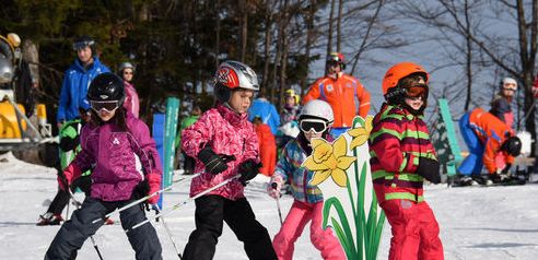 Kinder in bunten Skiklamotten üben sich auf einer verschneiten Piste im Skifahren, mit Erwachsenen und Bäumen im Hintergrund und einer großen gelben Blumendekoration im Vordergrund.