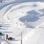 Kinder warten mit ihren Snowtubes am Fuße eines präparierten, verschneiten Tubing-Hügels, der von Zäunen und orangefarbenen Sicherheitsnetzen umgeben ist.